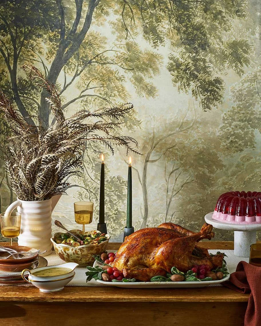 A Thanksgiving feast with a roasted turkey, cranberry dish, candles, and dessert on a rustic wooden table against a nature-themed wall mural.