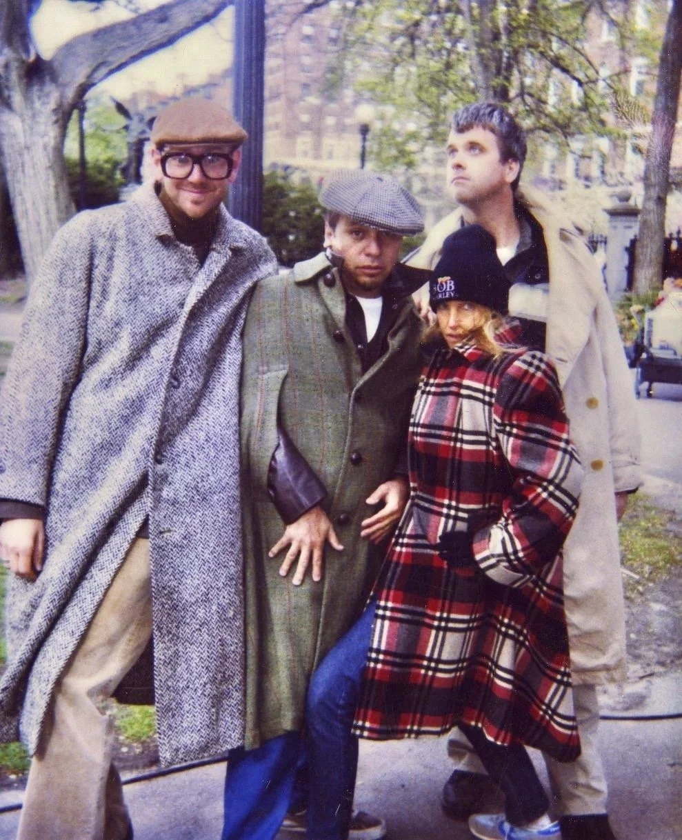 A group of four people outdoors in a park, wearing winter coats and hats, standing close together and posing for the camera.
