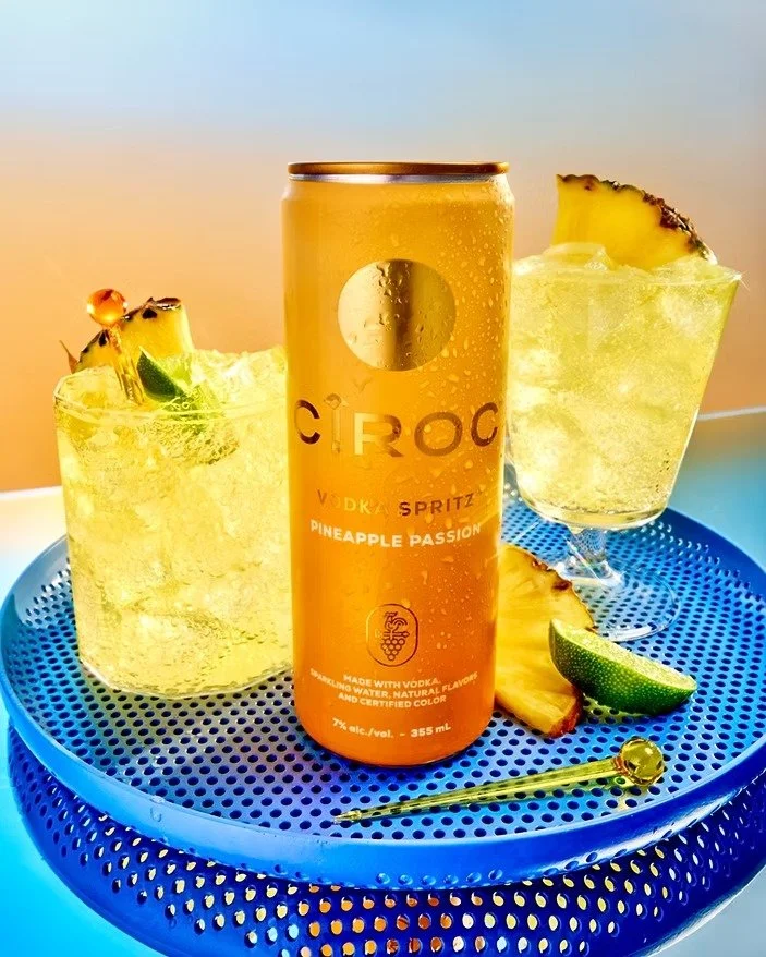 Two pineapple passion fruit cocktails with pineapple slices and lime wedges on a blue tray, with a canned vodka drink in the center.