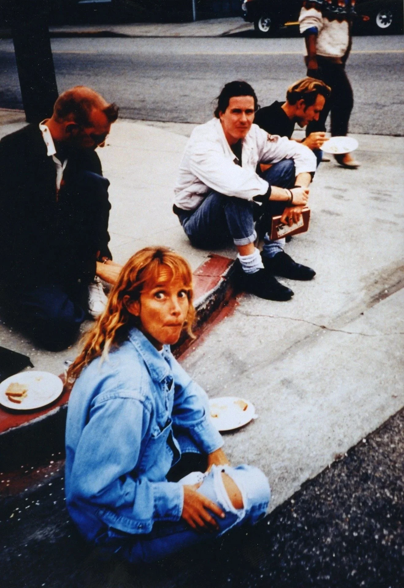 A group of people, including a woman with red hair wearing a blue denim jacket, sitting on a sidewalk outside, with some eating from plates. A man in a white jacket is squatting with a plate in his hand, and another person is sitting nearby. There ar