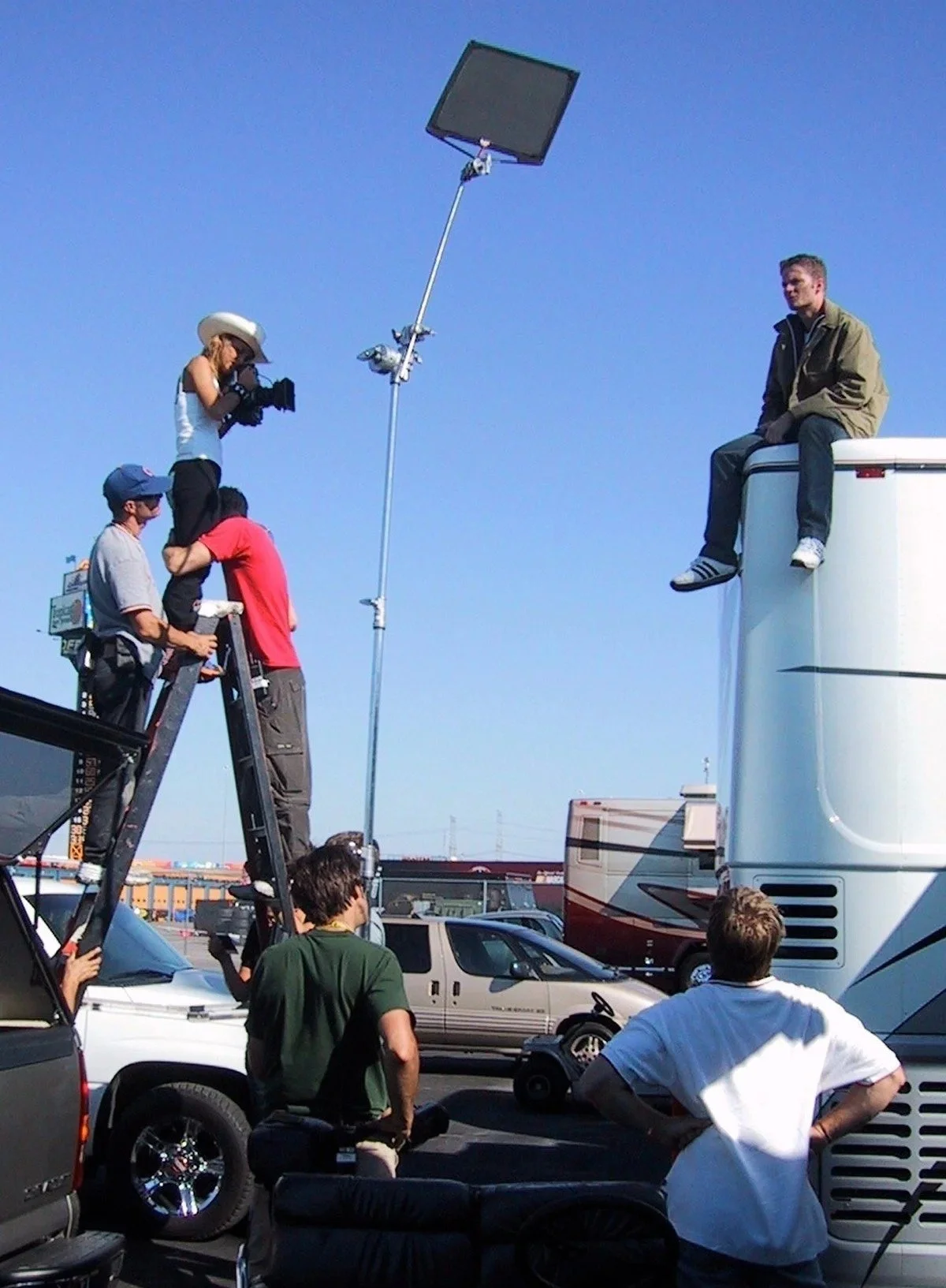 A film crew filming a man sitting on top of a large trailer. The crew is using a tall ladder, a camera crane, and other equipment, with several people watching or assisting. The scene is outdoors, in a parking lot under clear skies.