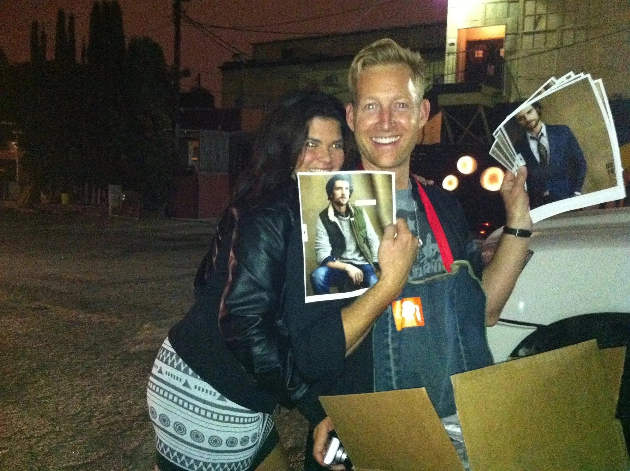Two people smiling at a nighttime outdoor event, holding photographs of a man in a leather jacket and a blue blazer, with one person holding a series of small round lights or decorations.