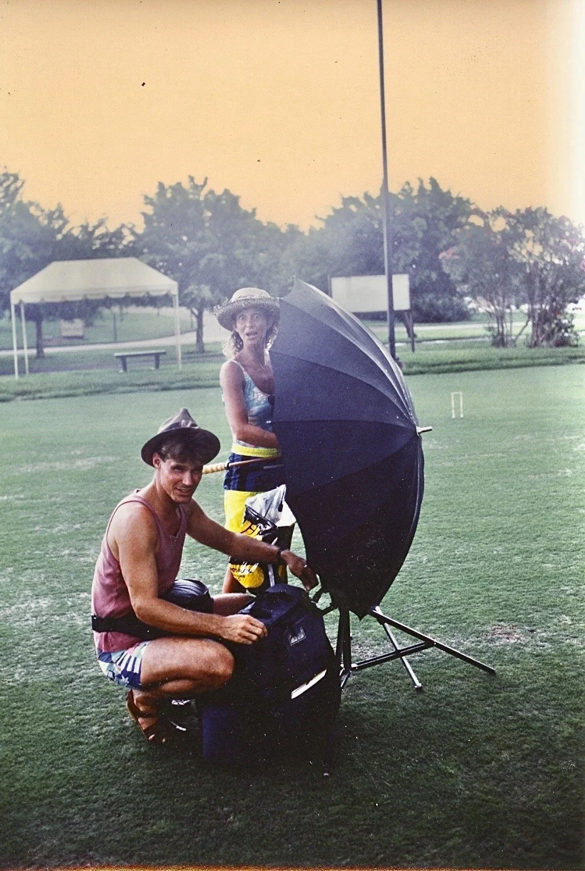 Two people in casual summer clothes and hats with an umbrella in an outdoor park with grass, trees, and a small tent in the background.