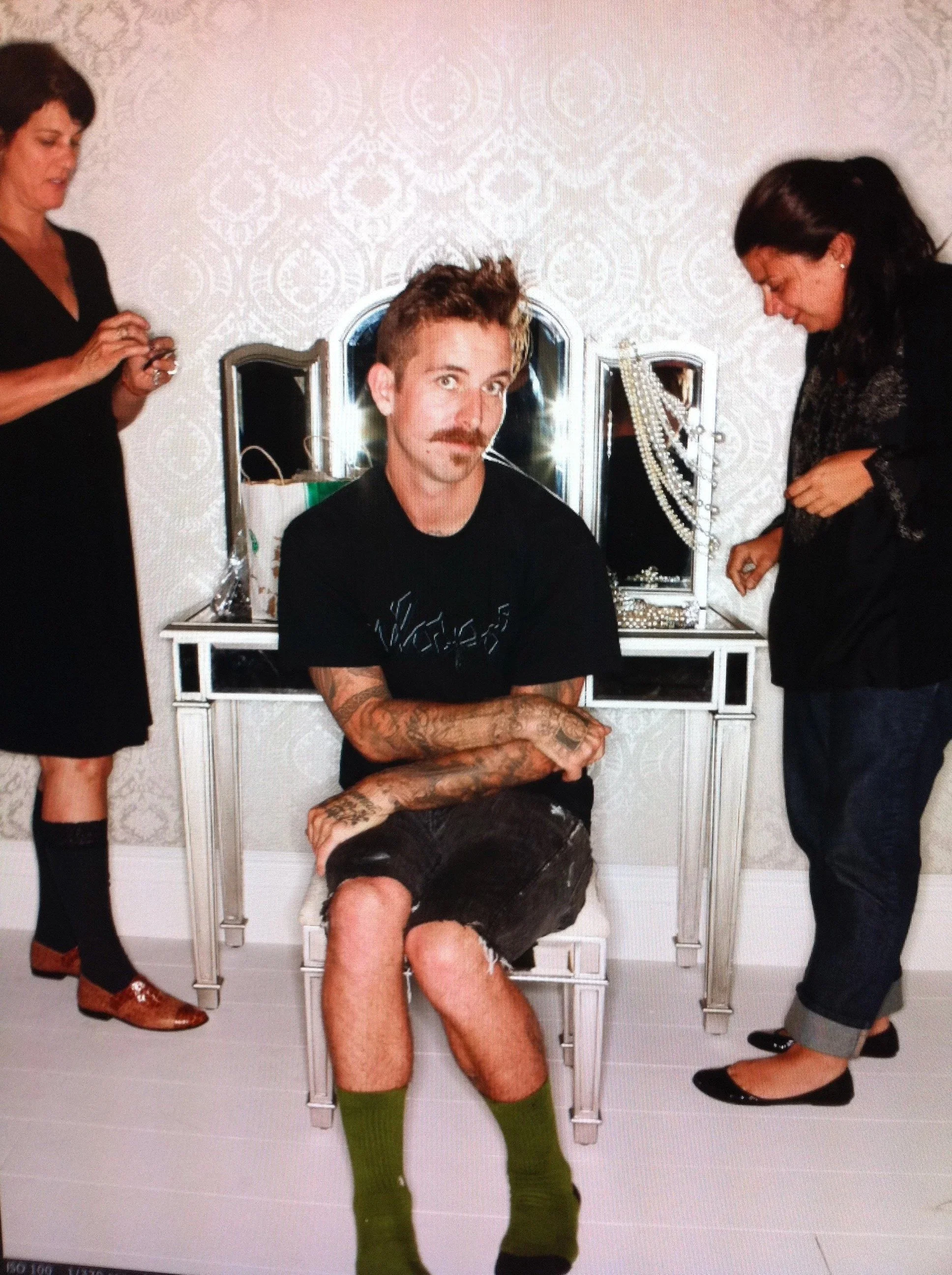 A young man with tattoos, a mustache, and spiky hair sitting on a white dressing table with his arms crossed, wearing black shorts and green knee-high socks. Two women, one on each side, are attending to him in a room with patterned wallpaper.