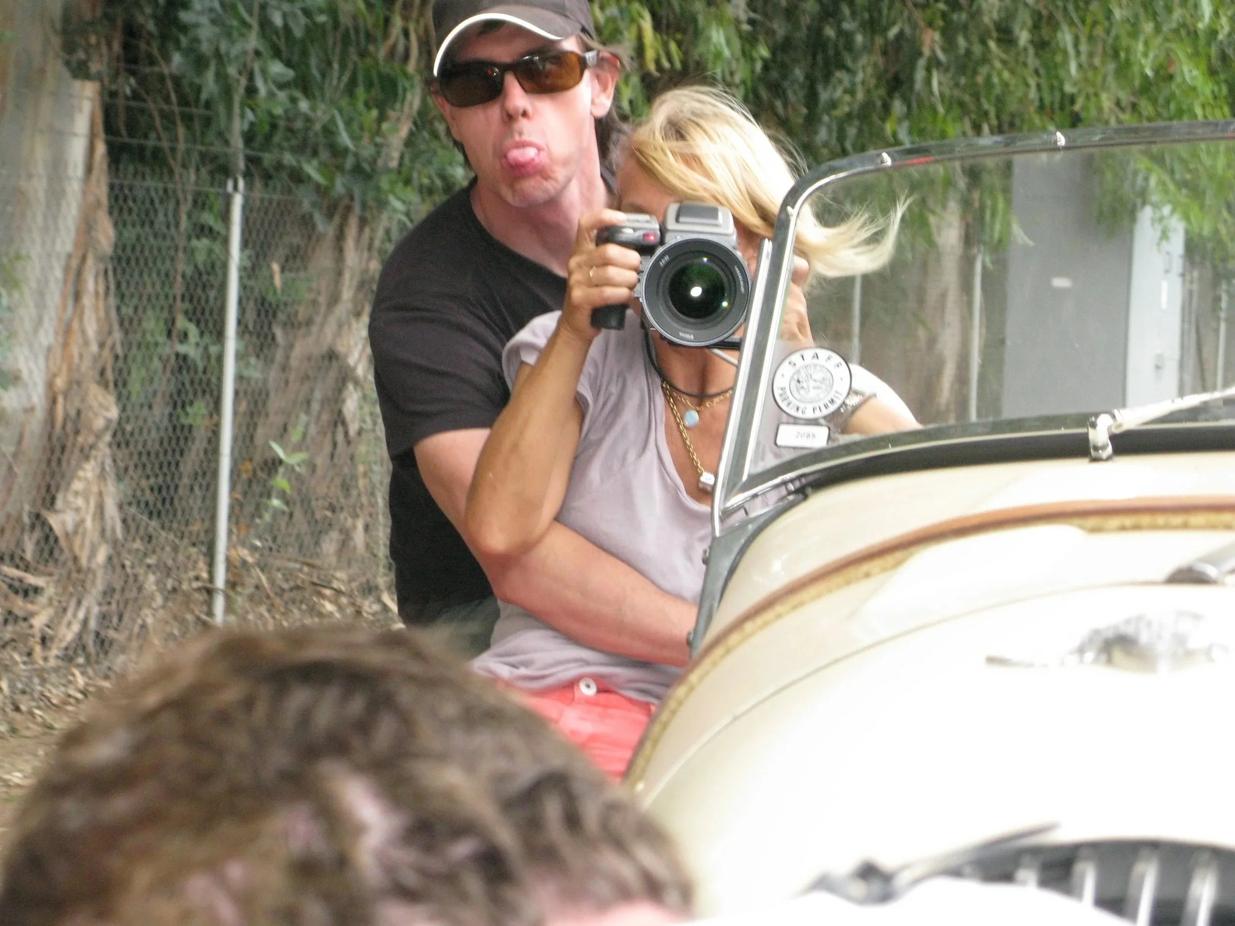 A man with dark hair wearing sunglasses and a black shirt sticking out his tongue, holding a camera, while a woman with blonde hair is sitting on a motorcycle with a windshield. They are outdoors with trees and a fence in the background.