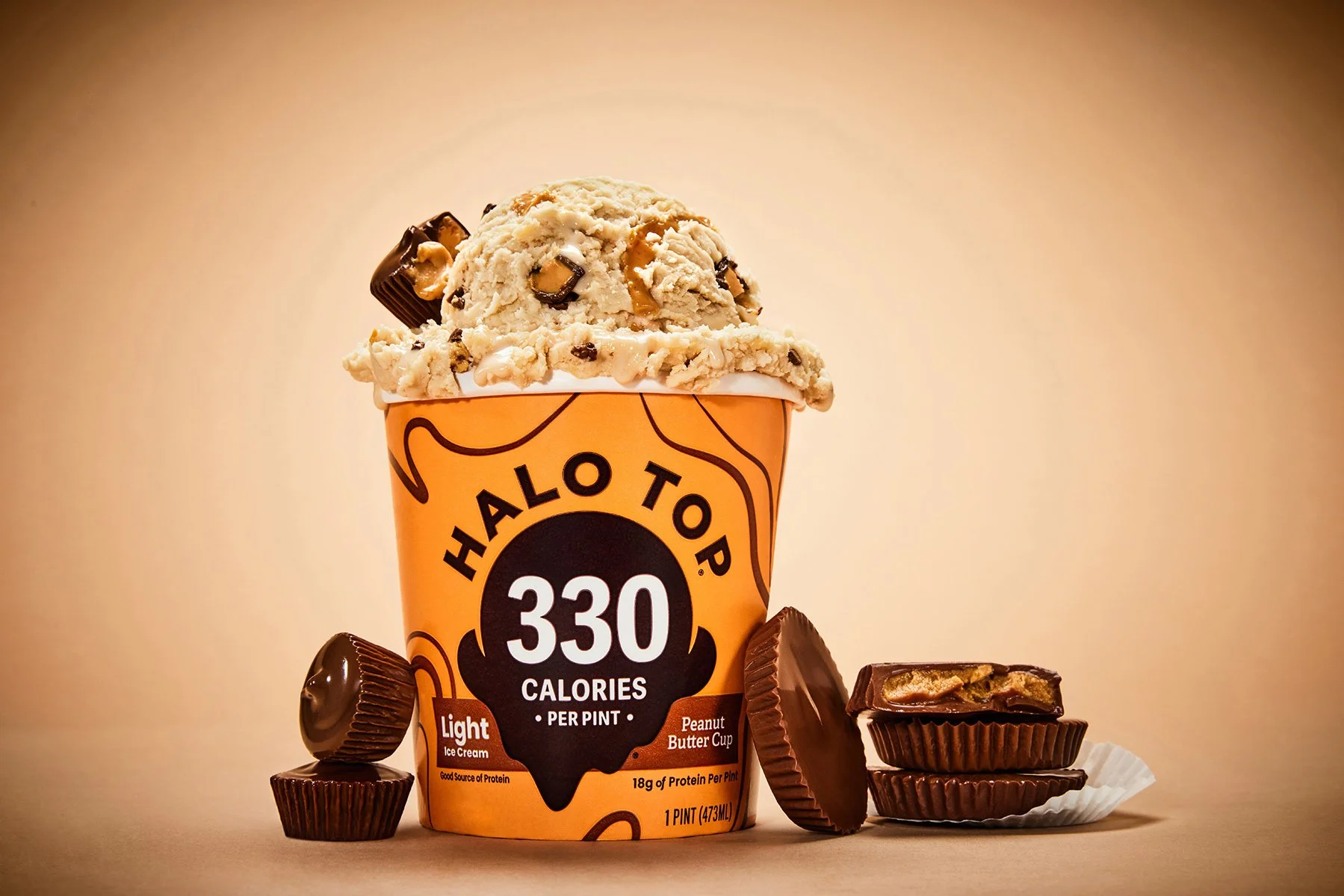 A pint of Halo Top cookie dough ice cream topped with cookie dough chunks and a piece of chocolate, surrounded by pieces of chocolate bars on a neutral background.