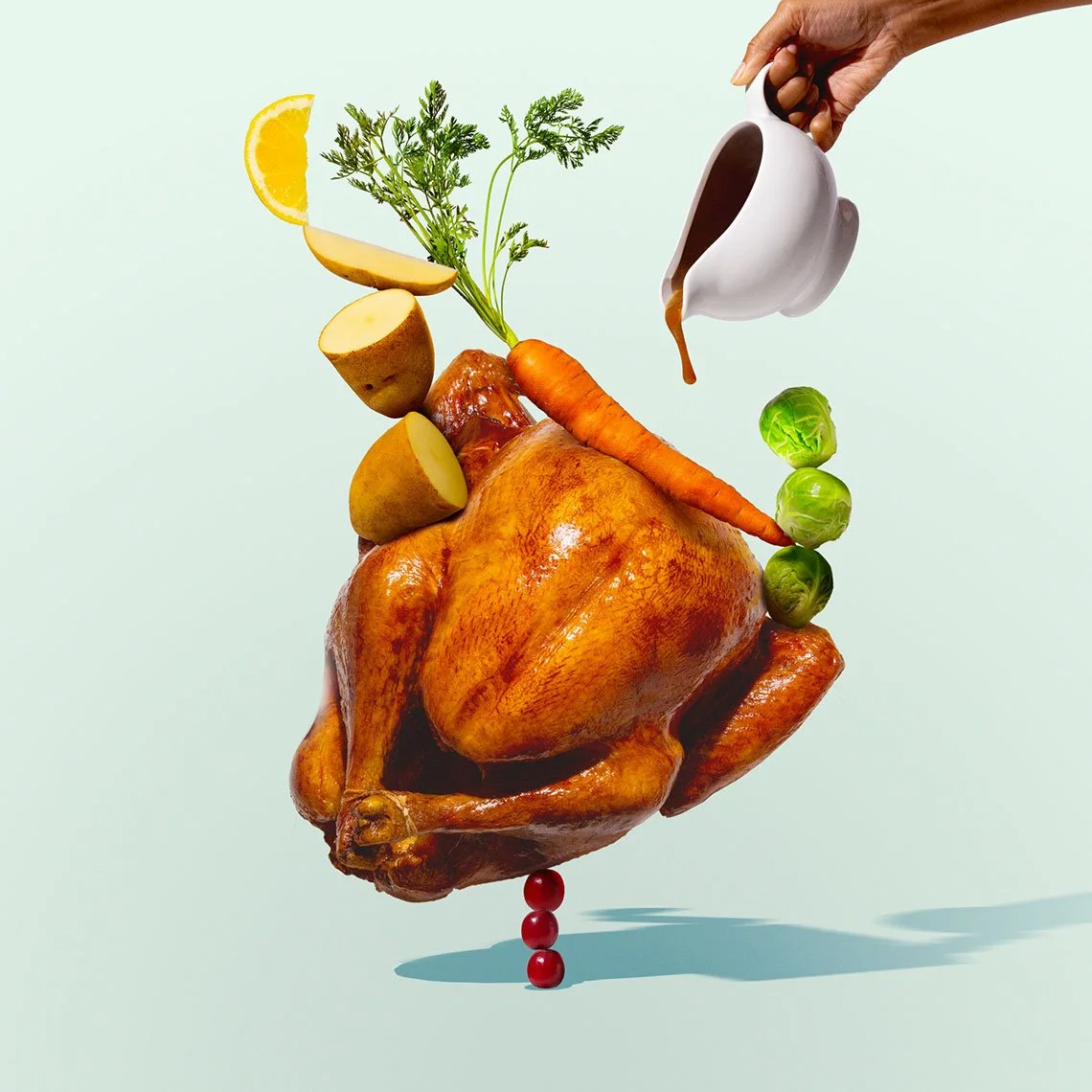 A roasted whole chicken with vegetables and herbs balanced with a lemon wedge, potatoes, carrot, Brussels sprouts, and parsley, with a hand pouring gravy from a white gravy boat.