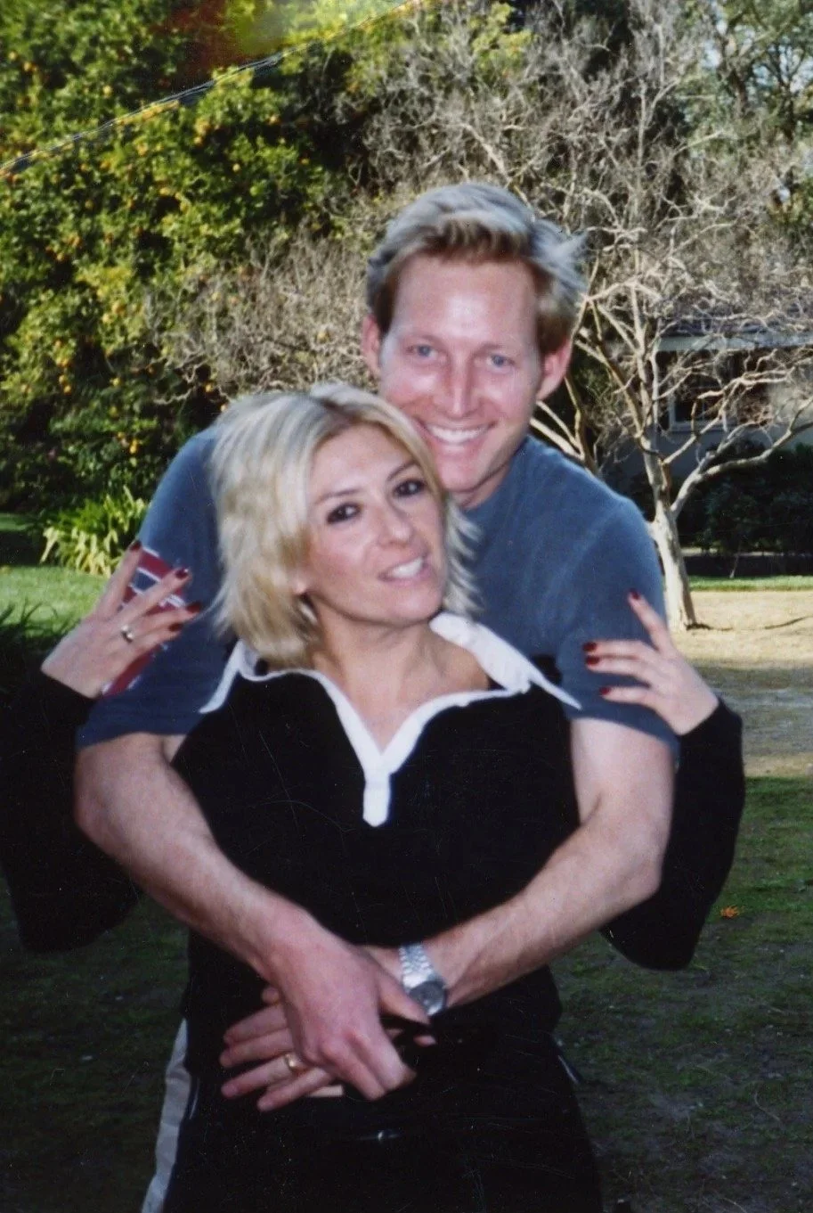 A smiling man and woman in a garden with trees, the man hugging the woman from behind.