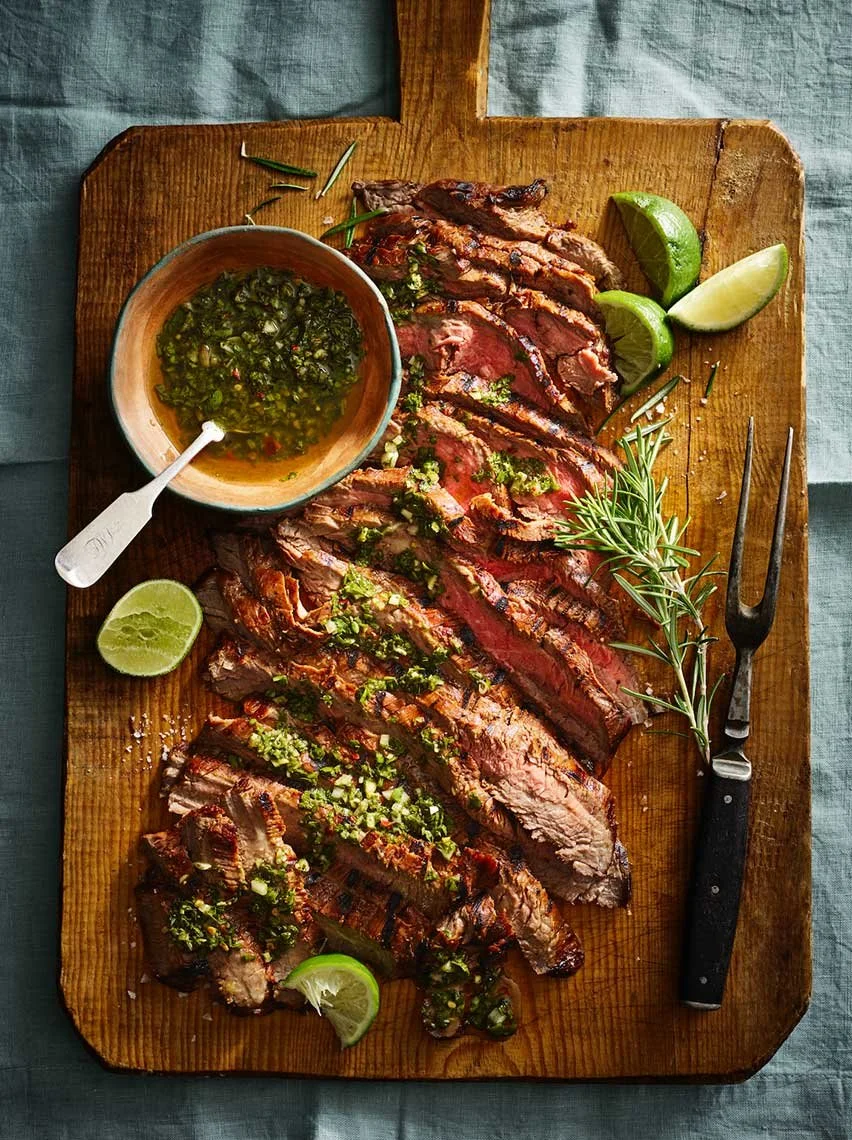 Sliced grilled steak topped with green herbs and chopped green onions on a wooden cutting board, with lime wedges, a sprig of rosemary, and a bowl of green sauce nearby.