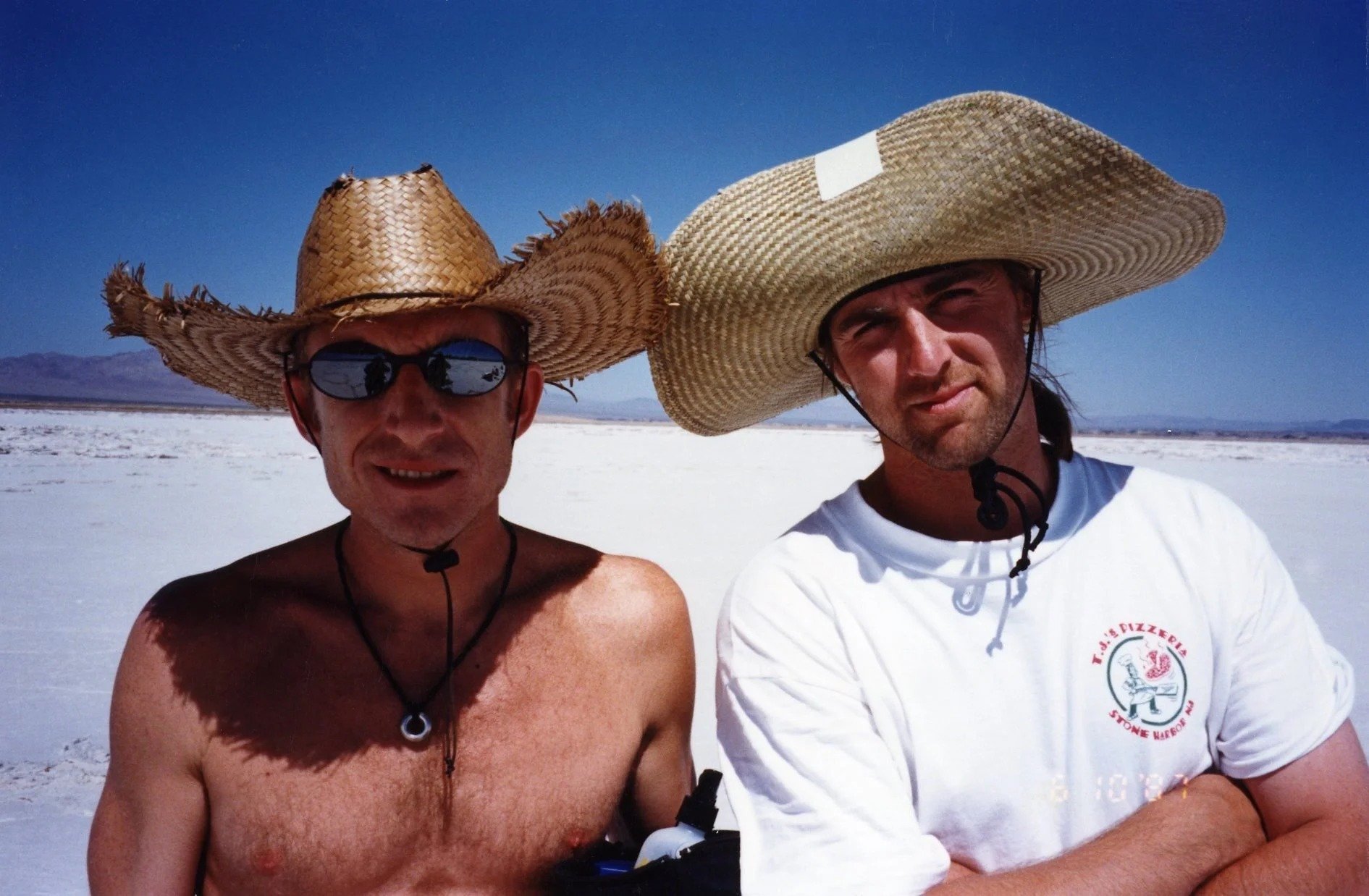 Two men wearing large straw hats, sunglasses, and casual clothing walking together on a salt flat.