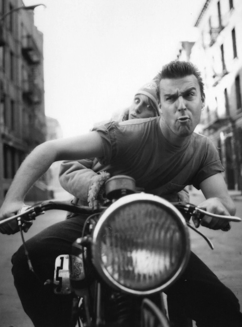 Two young men riding a motorcycle on an urban street, with one making a playful expression and the other looking ahead, surrounded by apartment buildings.