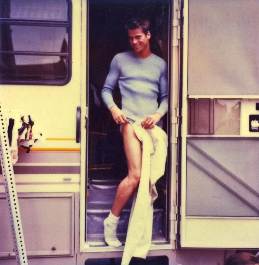 A man standing in the doorway of a camper van lifting his shirt to reveal his thigh and holding a blanket, smiling.
