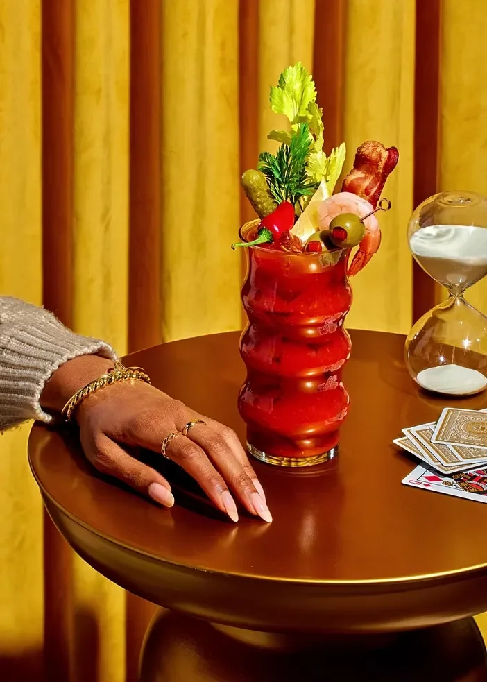 A hand with gold jewelry resting on a round wooden table next to a colorful cocktail garnished with celery, olives, and pepperoni, with an hourglass and a deck of cards nearby.