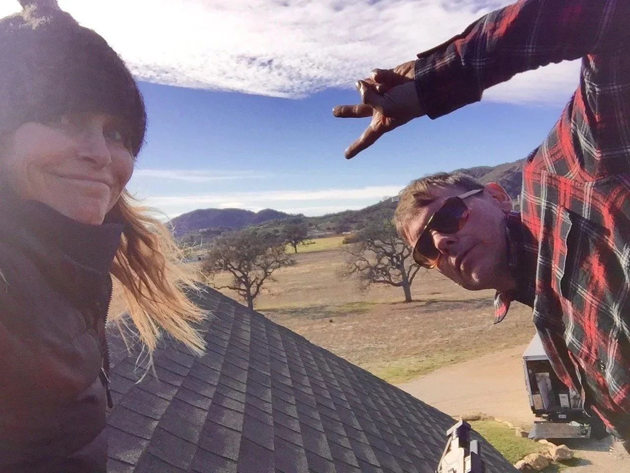 Two people taking a selfie on a roof with a rural landscape in the background, including trees, fields, and mountains, under a partly cloudy sky.