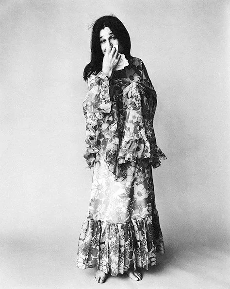 A woman standing in front of a plain background, wearing a floral patterned dress with ruffled sleeves and hem, covering her mouth with her left hand.