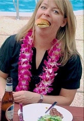 Woman with blonde hair wearing a black shirt and pink lei, holding a hot dog in her mouth at a poolside gathering, with a drink and food on the table.