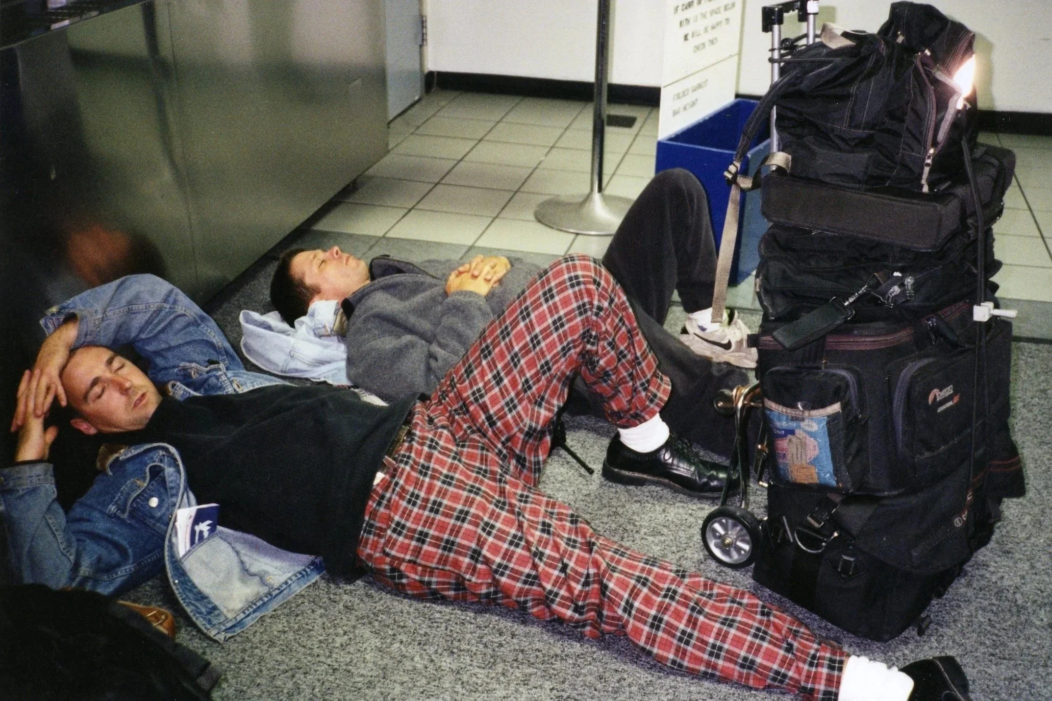 Three men are lying on the floor in an airport or train station, appearing to be asleep. They are surrounded by luggage, including backpacks and a stroller, and are positioned near a metal counter and a sign with instructions.