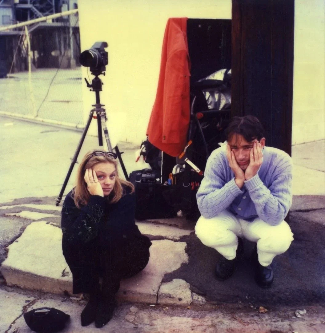 A woman and a man crouching on a cracked sidewalk, with camera equipment and a tripod nearby, in front of a yellow wall and an open door.