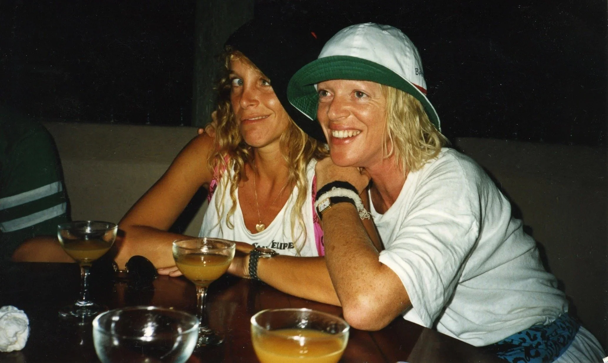 Two women sitting at a table with drinks, smiling and enjoying each other's company in a dimly lit setting.
