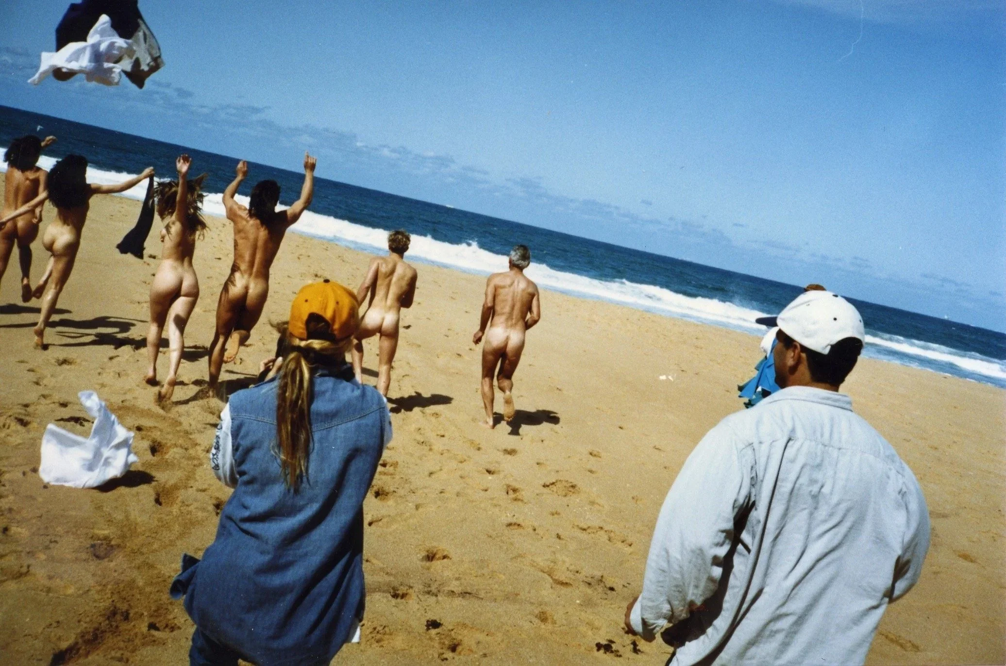 People, mostly nude, running on a sandy beach toward the ocean, with a blue sky and some clouds overhead.