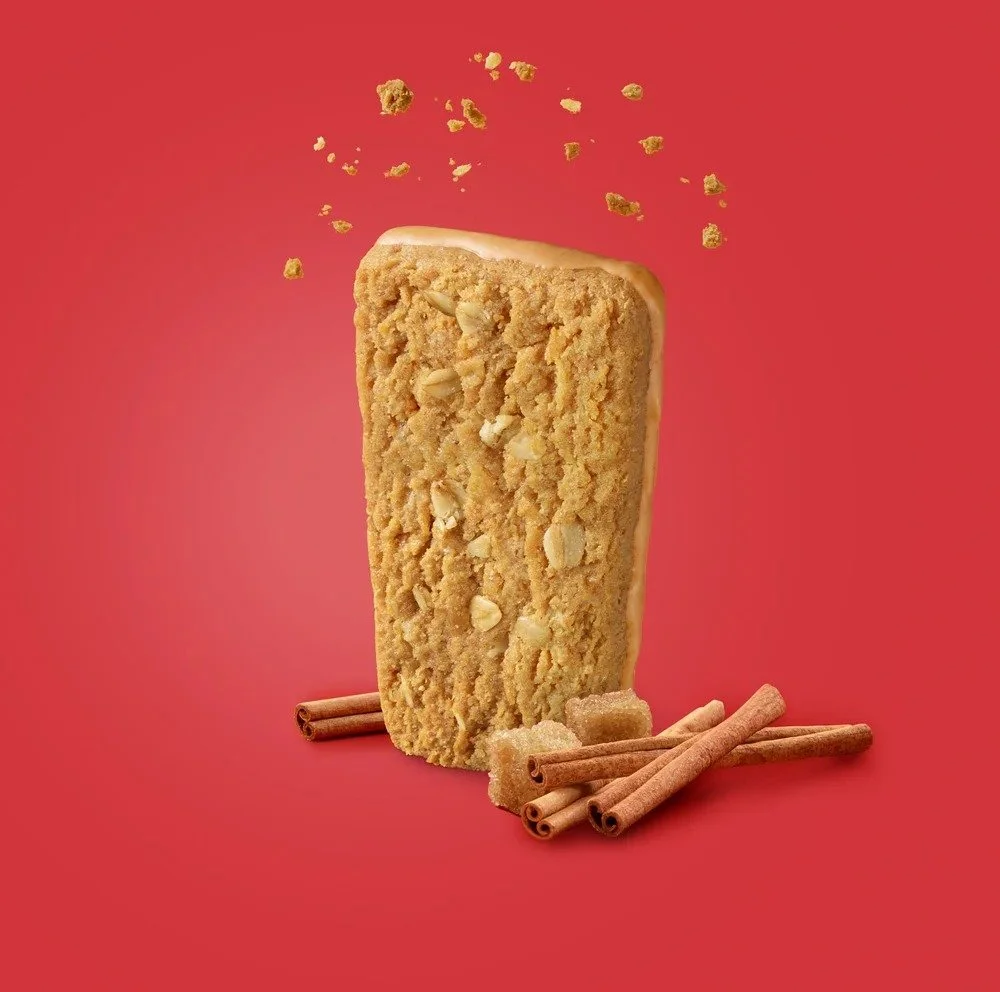 A large cookie with white chocolate chunks standing upright on a red background, surrounded by cinnamon sticks and sugar cubes, with cookie crumbs floating around.