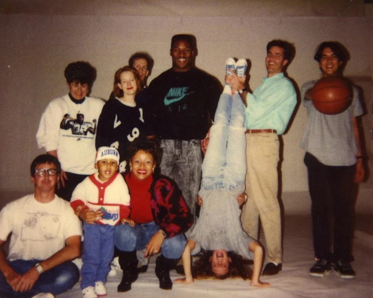 A group of ten diverse people posing together indoors, some making playful gestures, with one person holding a basketball and another person flipping upside down on the floor.