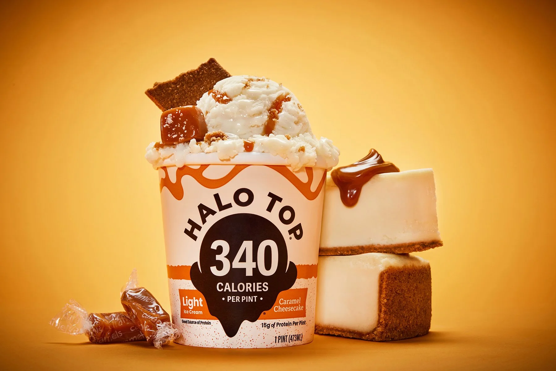 A pint of Halo Top ice cream with caramel cheesecake flavor topped with caramel sauce, with caramel candies and slices of cheesecake nearby.