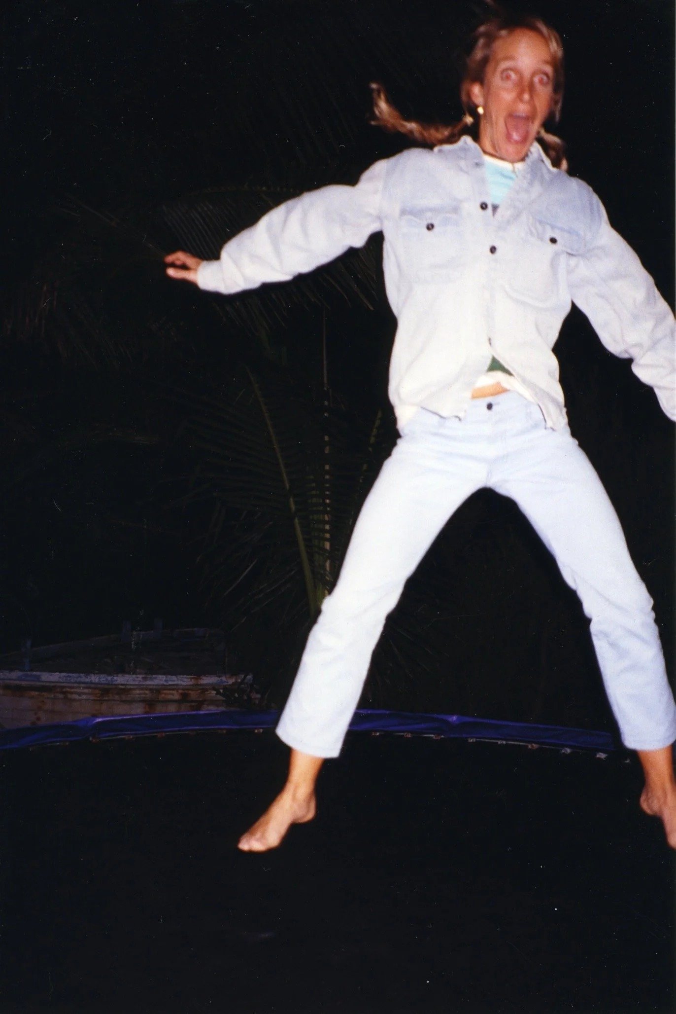 A woman with long red hair in pigtails, wearing a white jacket and white pants, jumping or balancing on a trampoline at night with a dark sky background.