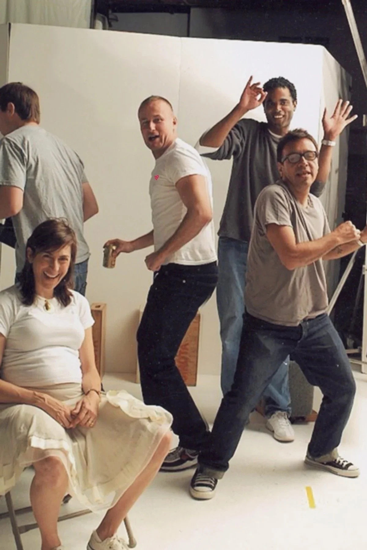A group of five people in a photo studio, with six of them visible, including a woman sitting on a chair smiling, and four men standing behind her dancing and making playful gestures. One man is holding a camera or similar object and some are wearing