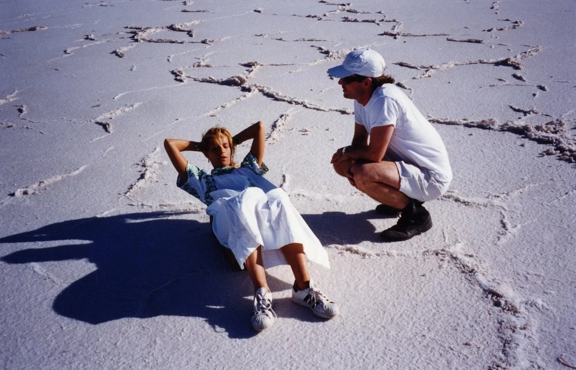 A person is lying on the salt flats, possibly the Salar de Uyuni in Bolivia, with their hands behind their head. Another person is squatting nearby, looking at them. The ground is covered in white salt with visible cracks forming patterns.
