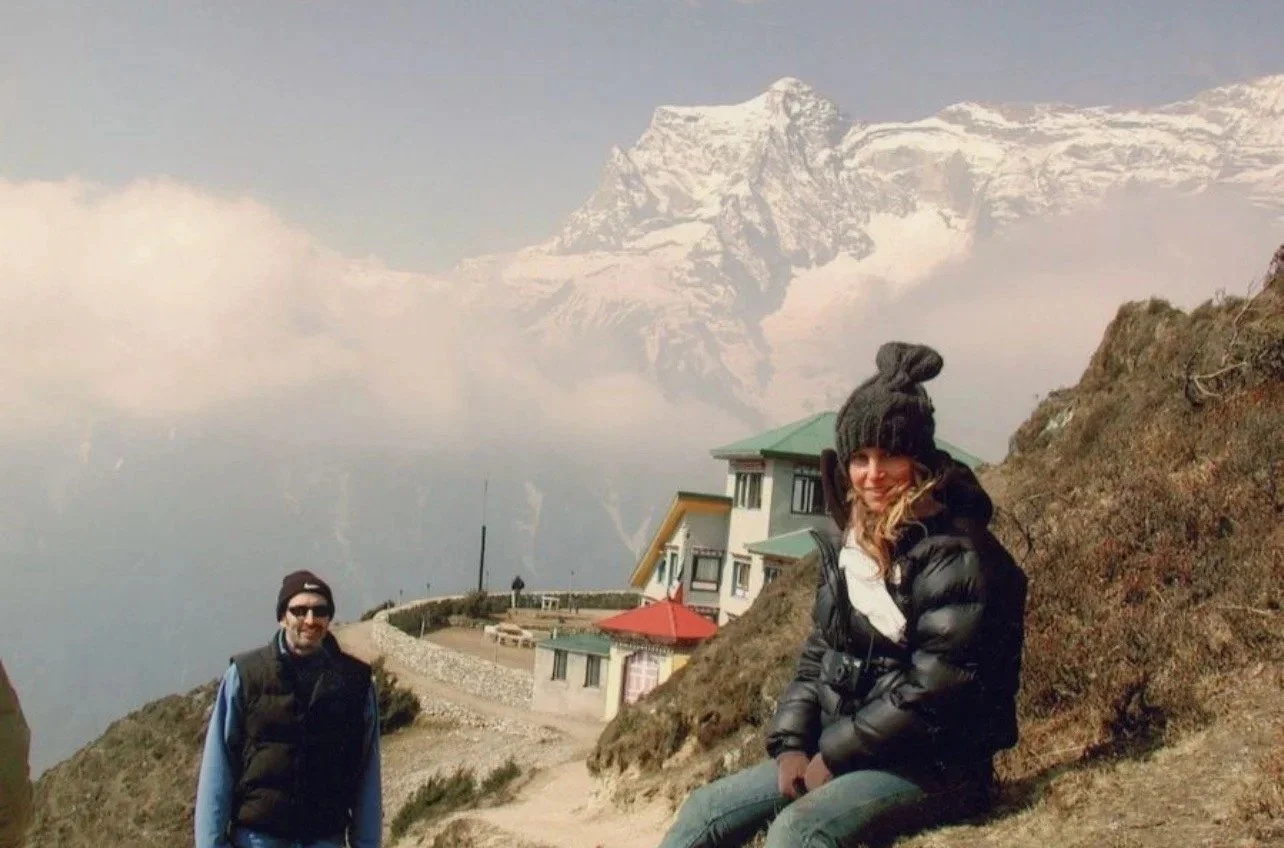 Two people enjoying outdoor mountain scenery near a building with a snow-capped peak in the background.