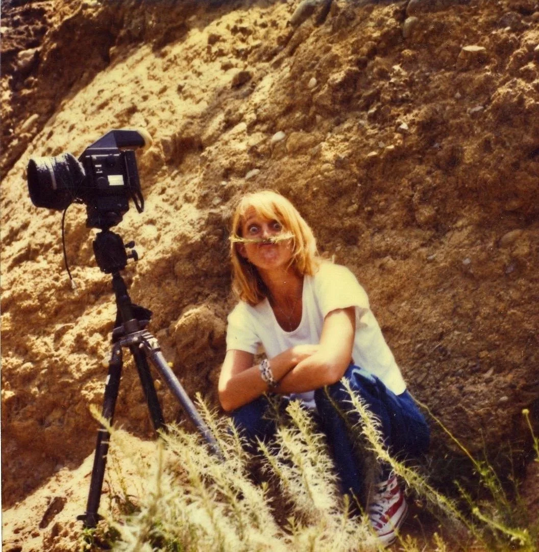 A woman with red hair wearing a white t-shirt sitting on the ground in front of a camera on a tripod, with a nose implant, surrounded by sandy terrain and dry grass.