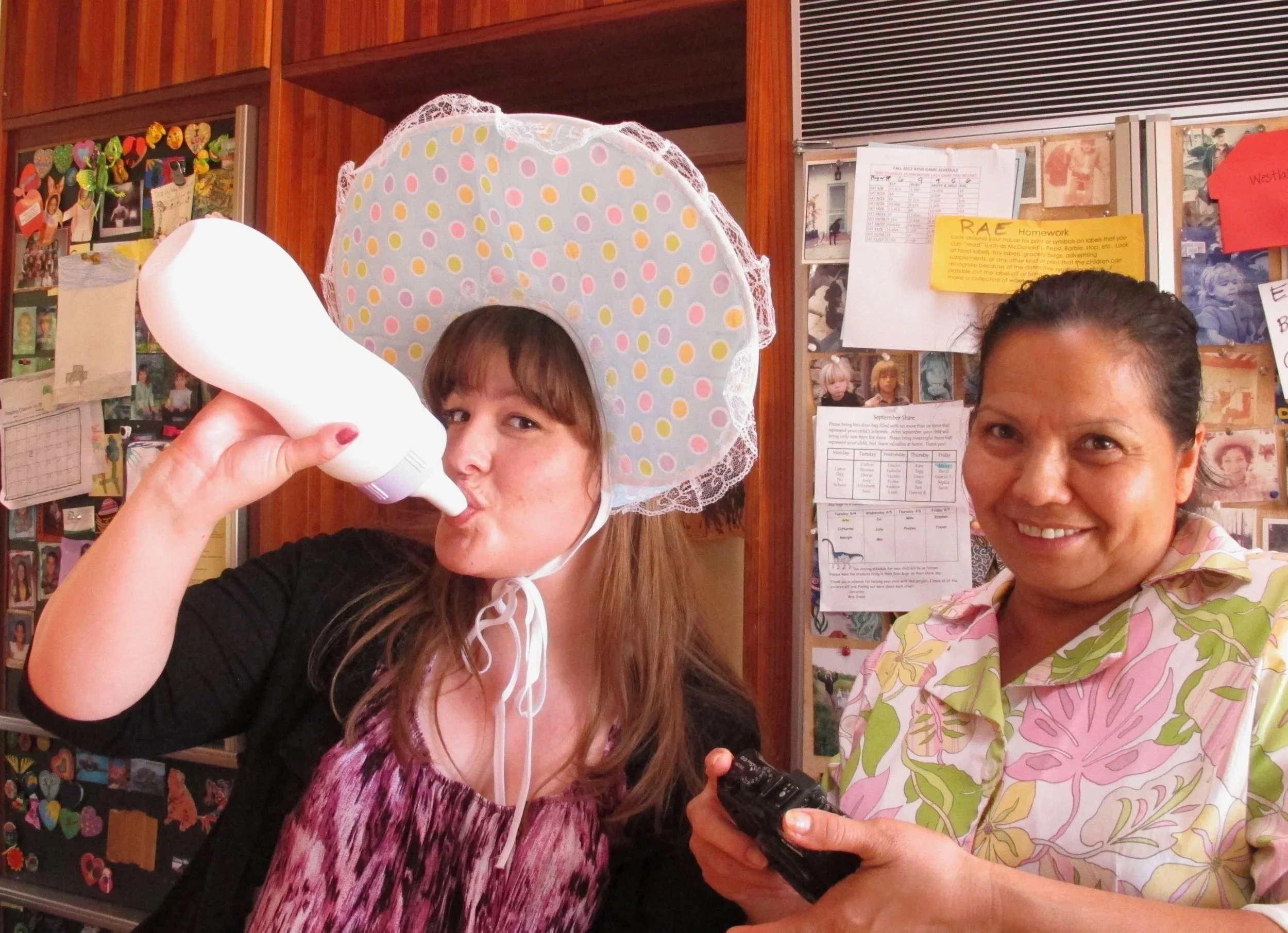 Two women smiling and posing for a photo indoors. One woman is wearing a large, polka-dotted, bonnet-like hat and holding a hairdryer near her face. The other woman, dressed in a floral top, is partially visible and smiling. The background shows bull