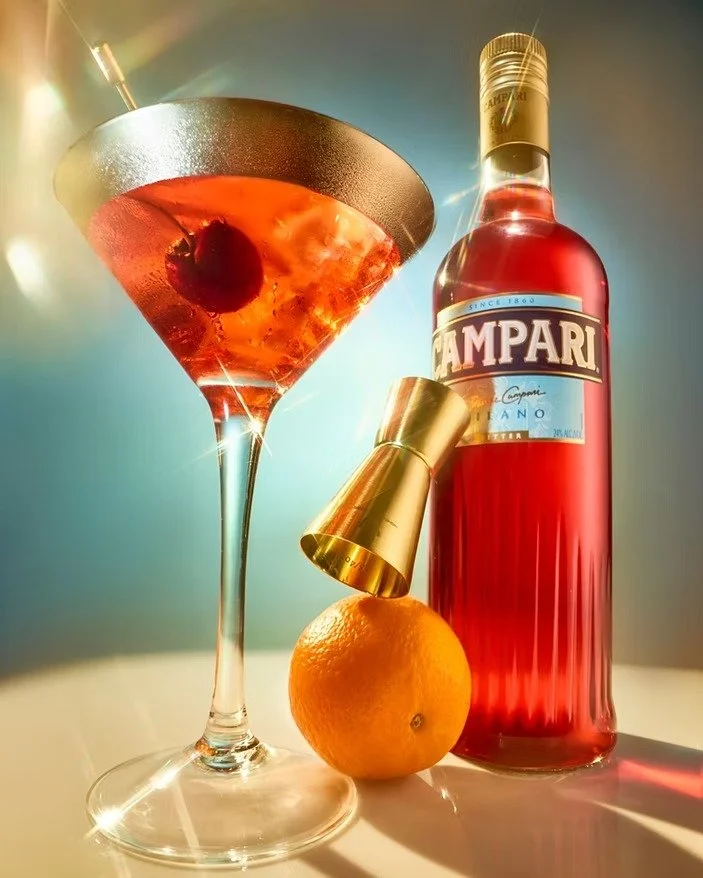 A cocktail glass with a cherry garnish, a bottle of Campari, an orange, and a gold jigger on a white surface with colorful lighting.