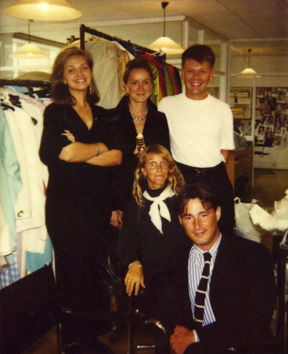Group of five people in a clothing store, posing for a photo. Four are standing, one is sitting in front. Racks of clothes and a wall of photos are in the background.