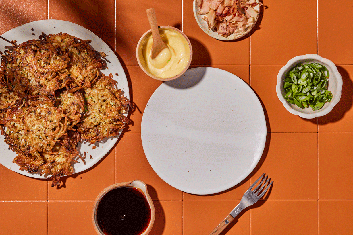 A table set with a plate of crispy onion rings, a cup of mayonnaise, a small bowl of chopped ham, a small bowl of chopped green onions, a cup of soy sauce, an empty white plate, a fork, and a glass of soy sauce on an orange tiled surface.