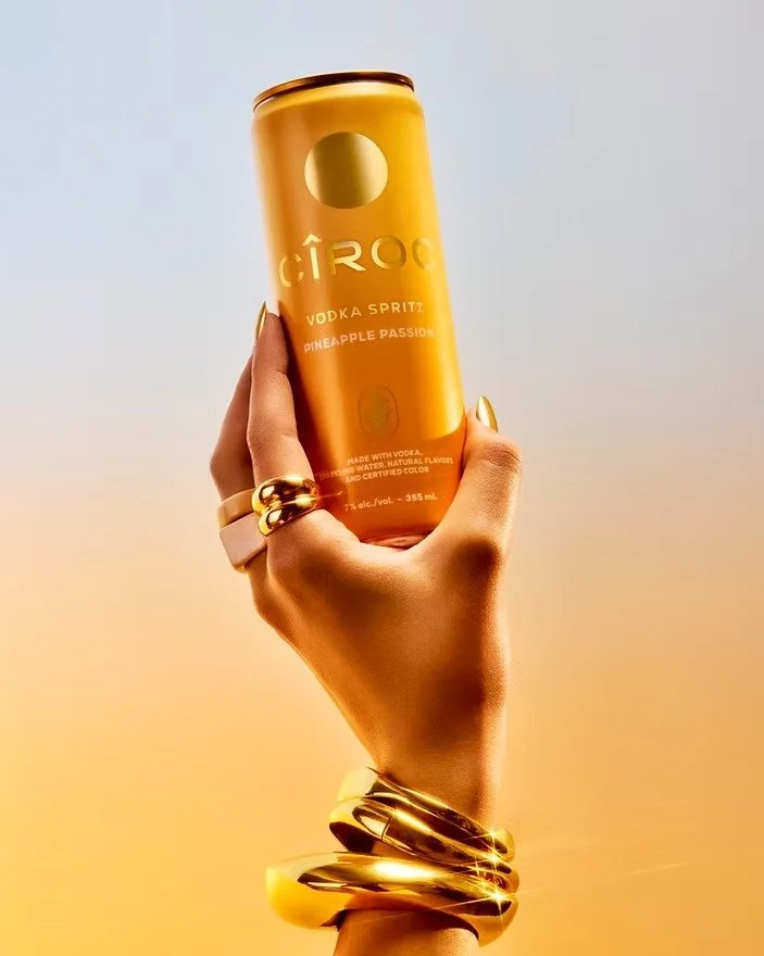 A hand with gold rings and bracelets holding a yellow can of Cîroc Pineapple Passion vodka spritz against a sunset background.