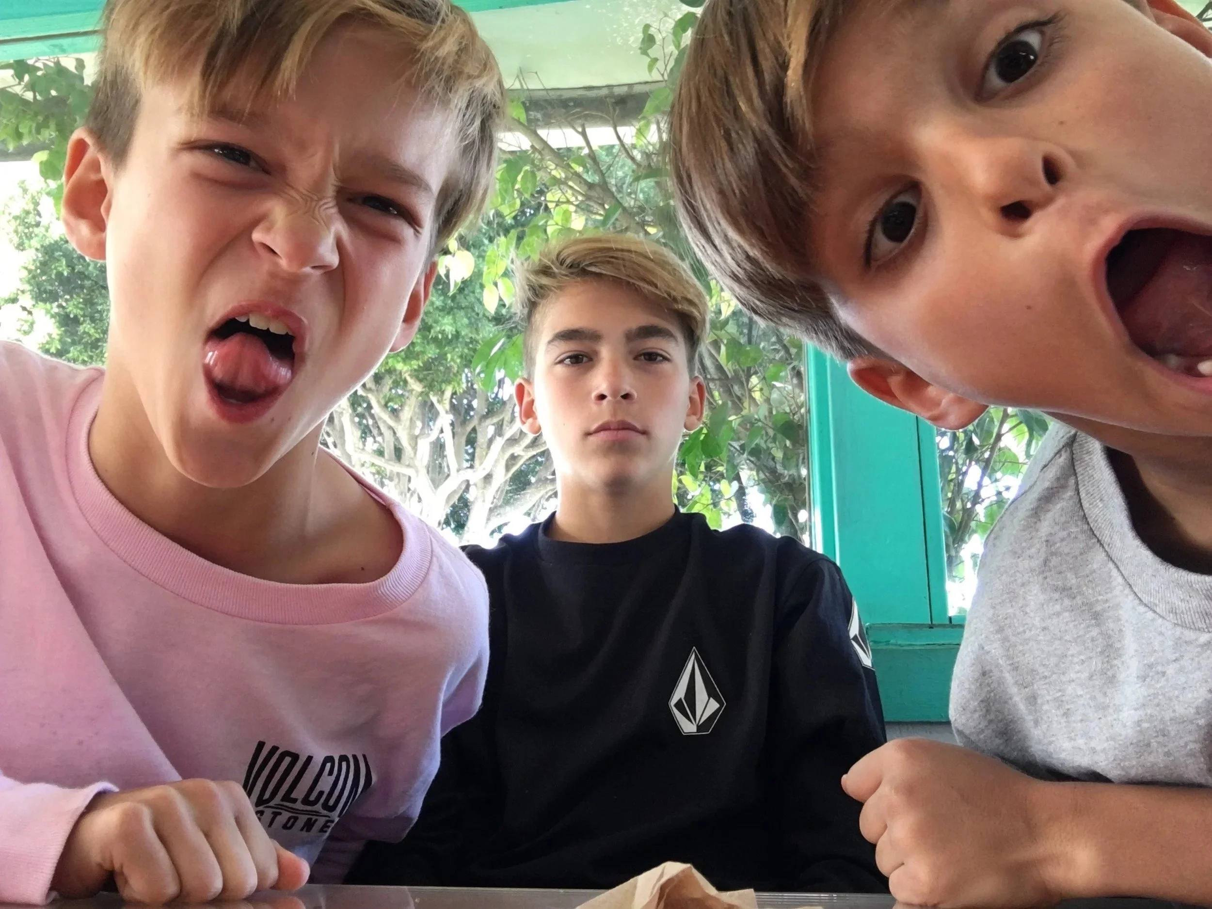 Three boys are close to the camera, with two on the sides and one in the middle, all making different facial expressions. The boy on the left has an angry or playful expression, sticking out his tongue and furrowing his brows. The boy on the right ha
