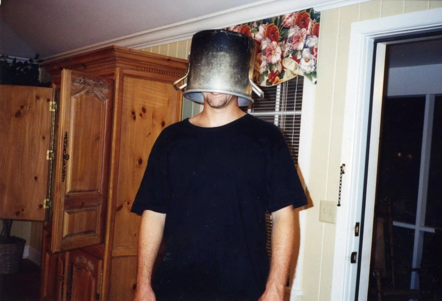 A person standing indoors with a large black metal bucket covering their head. The room features wooden cabinets, a floral-patterned curtain, and a partially open sliding glass door at night.