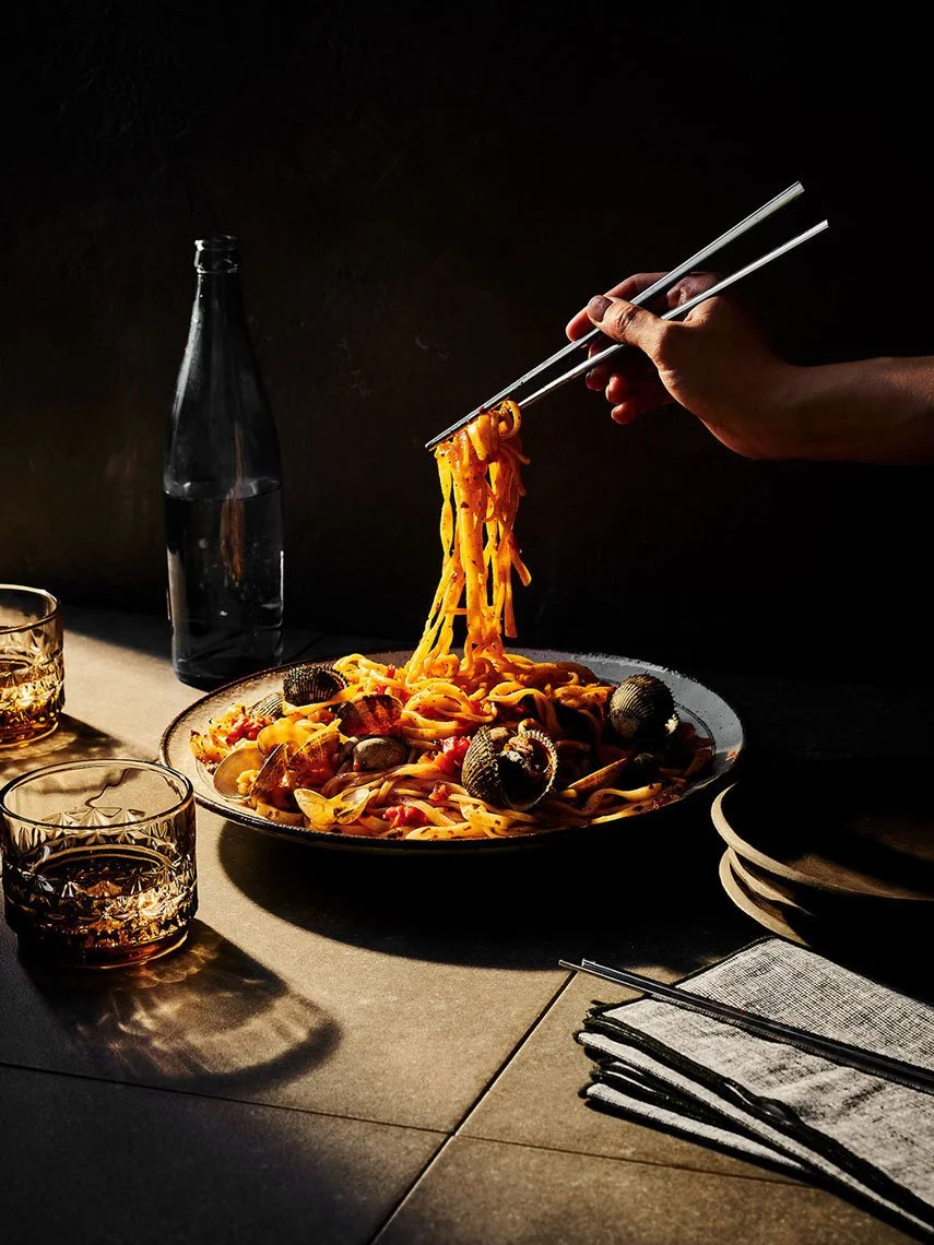Person using chopsticks to lift pasta with clams from a plate, on a table with glasses of dark beverage, a water bottle, and cloth napkin, against a dark background.