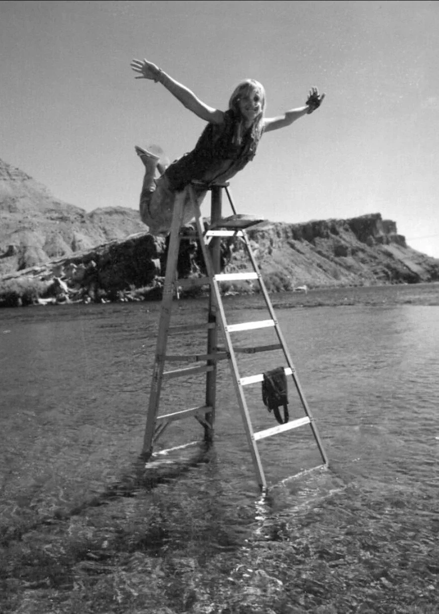 A woman standing on a ladder in the water at the beach, with arms outstretched, and cliffs in the background.