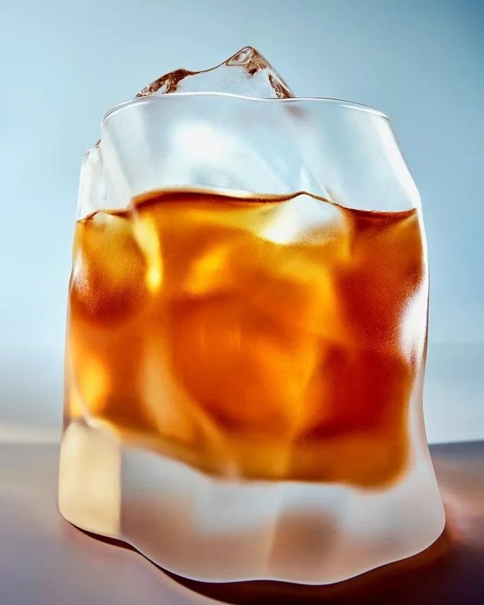 Close-up of a glass containing brown iced coffee with ice cubes, viewed against a light background.