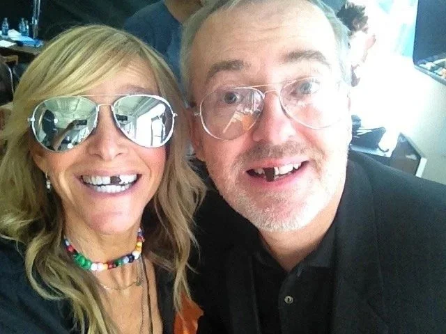 A woman and a man taking a selfie indoors. The woman is wearing reflective aviator sunglasses, has blonde hair, and is smiling. The man is wearing clear glasses, has short gray hair, a beard, and is also smiling.
