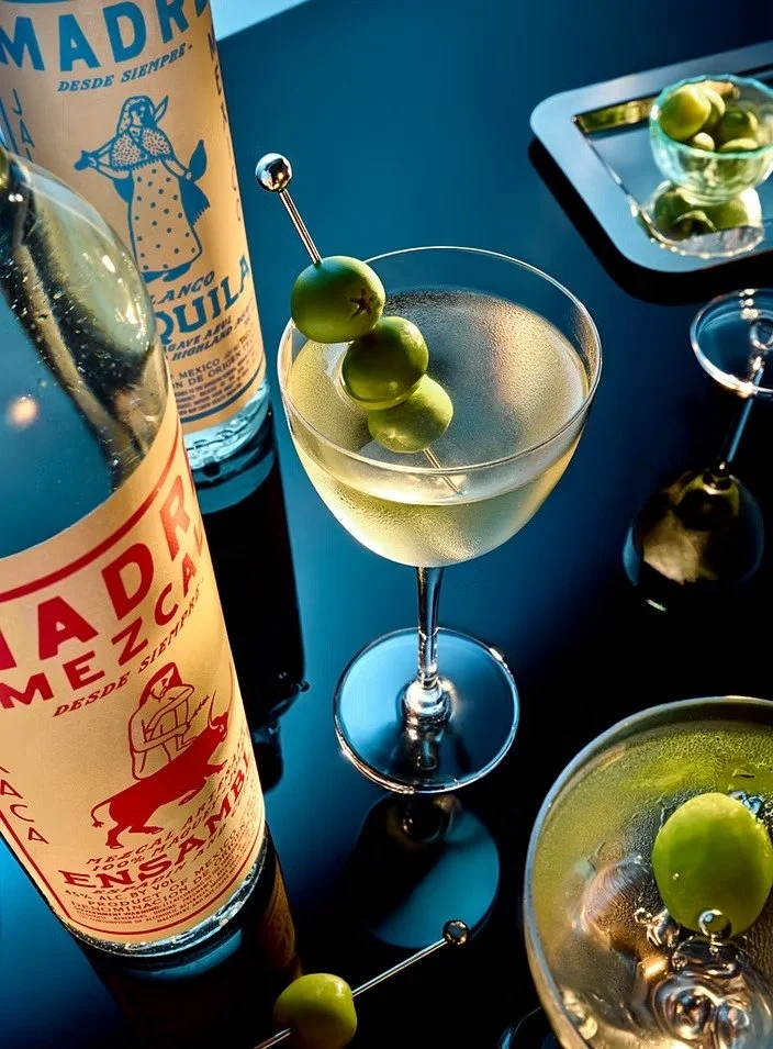 Martini glass with a green olive skewer, a bottle of tequila, a glass of olive garnished drink, and condiments on a dark table.