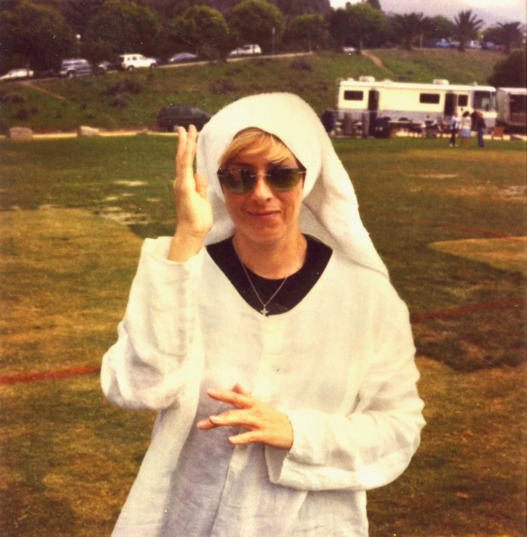 A woman wearing sunglasses, a white knit hat, and a white hoodie, standing outdoors on a grassy field with RVs and parked cars in the background.