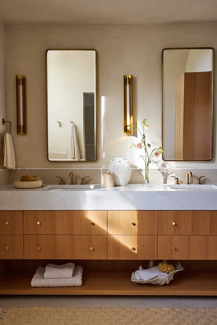 Modern bathroom vanity with two sinks, wooden cabinetry, and two tall mirrors. Decorative items include a white vase with flowers, towels, and candles. Wall-mounted lights provide warm lighting.