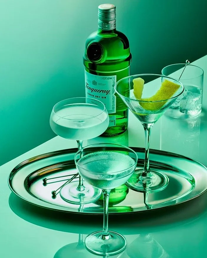A bottle of Tanqueray gin, a cocktail shaker, and three cocktail glasses with drinks, garnished with lemon peels, placed on a silver tray with cocktail picks.