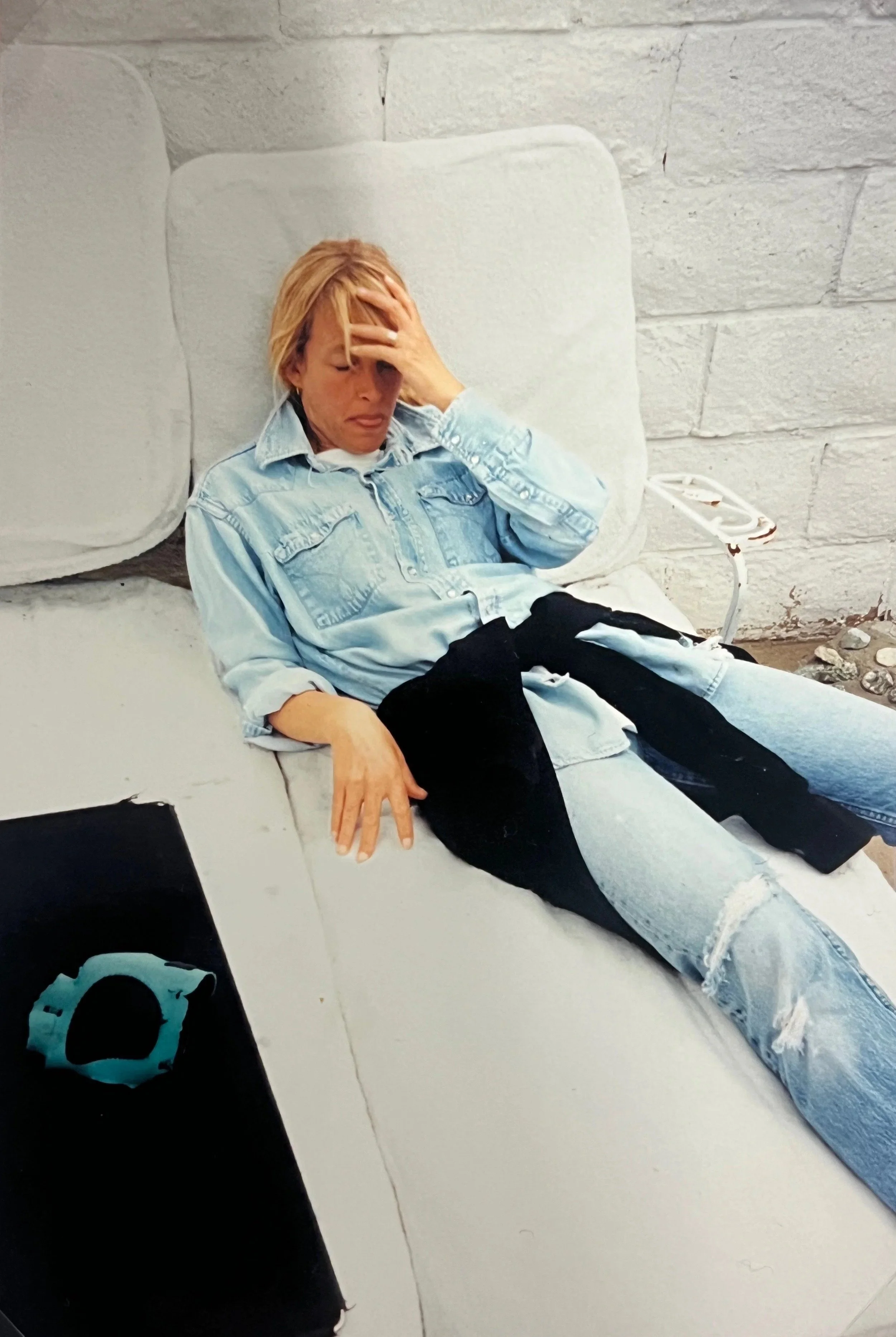 A woman sitting on a white bench with her eyes closed, head resting on her right hand, in front of a white brick wall.