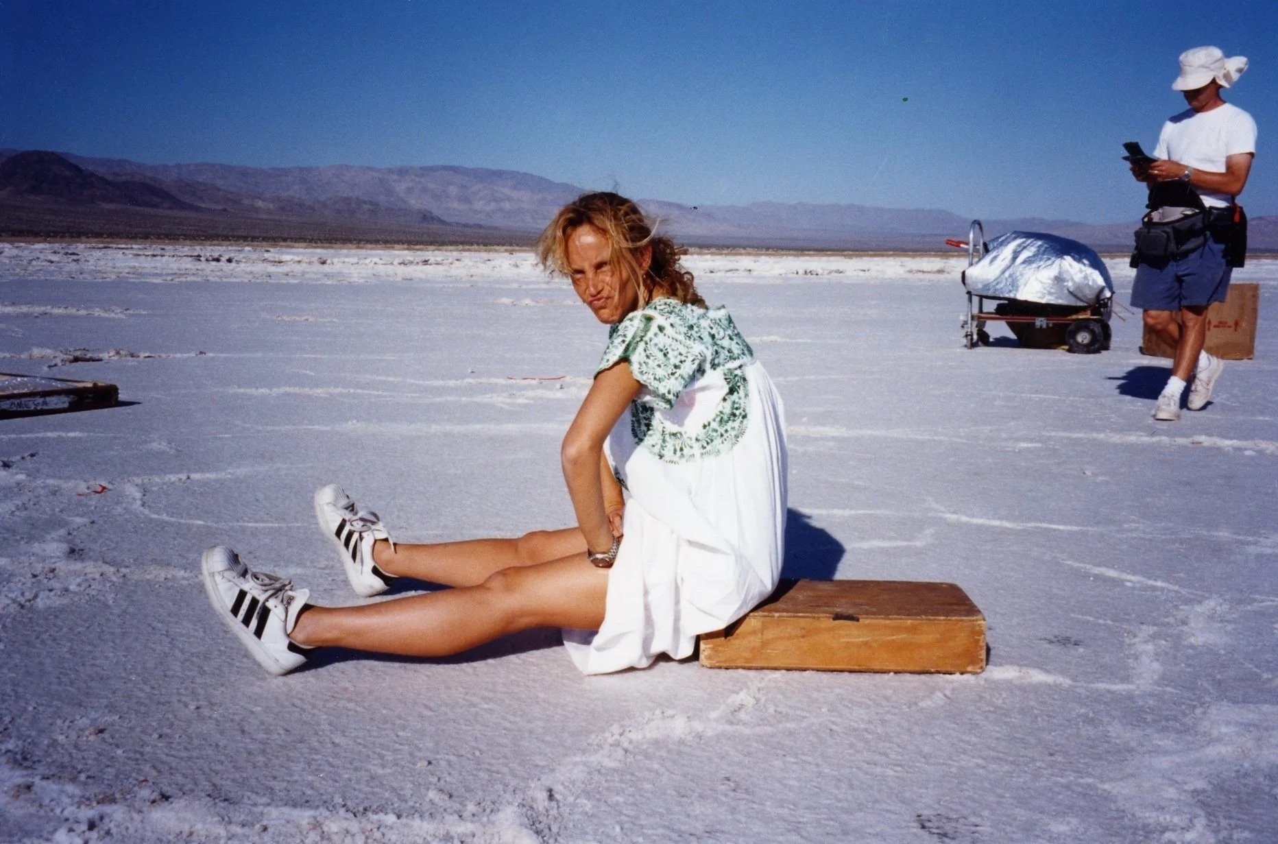 A woman in white Adidas sneakers and a white dress is sitting on a wooden bench, stretching her legs on a salt flat under a clear blue sky. In the background, a person wearing a white hat, white t-shirt, blue shorts, and sunglasses is standing, looki