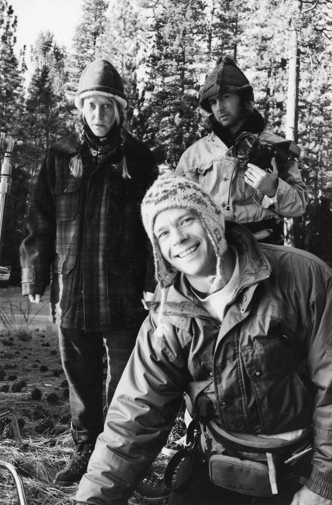 Black and white photo of three people outdoors in a forest. The person on the left is a woman with braided hair, wearing a hat, jacket, and a bandana. The person on the right is a man with a camera, wearing a hat and jacket. The person in the front i